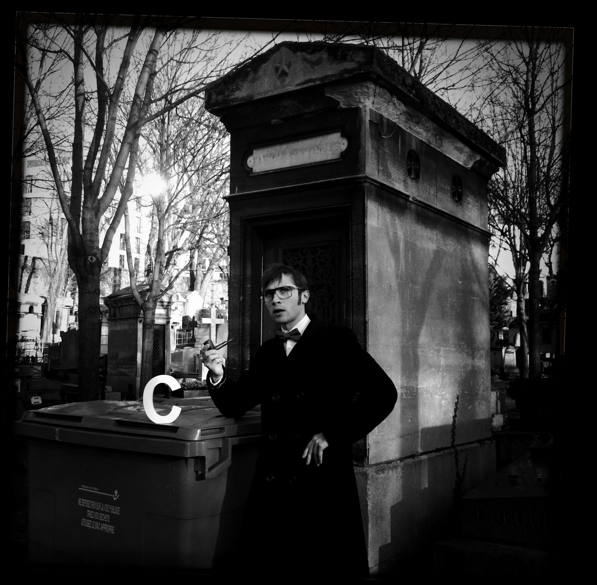 Caspar Schjelbred in PLAN C, in a long dark coat and bow tie holding a pipe, standing by a tomb in a Paris cemetery beside a white letter C